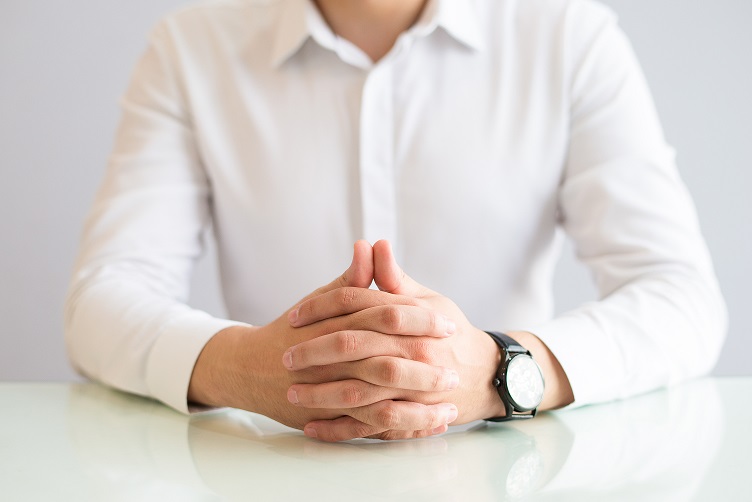 Closeup of man sitting at table with his hands clasped. Male person wearing shirt and watch. Concentration or waiting concept. Cropped front view.